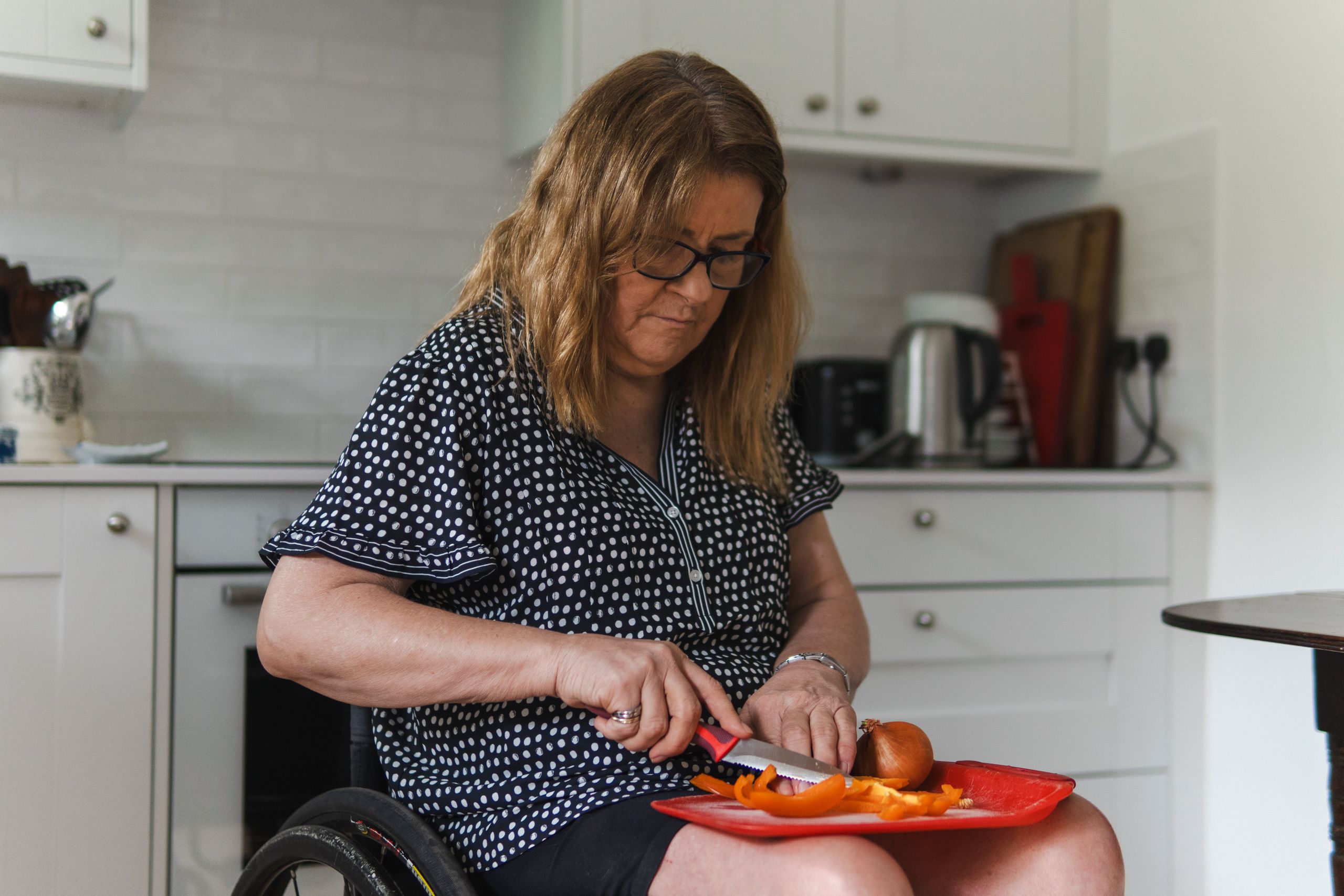 A woman in a wheelchair cutting vegetables on a chopping board in a kitchen.