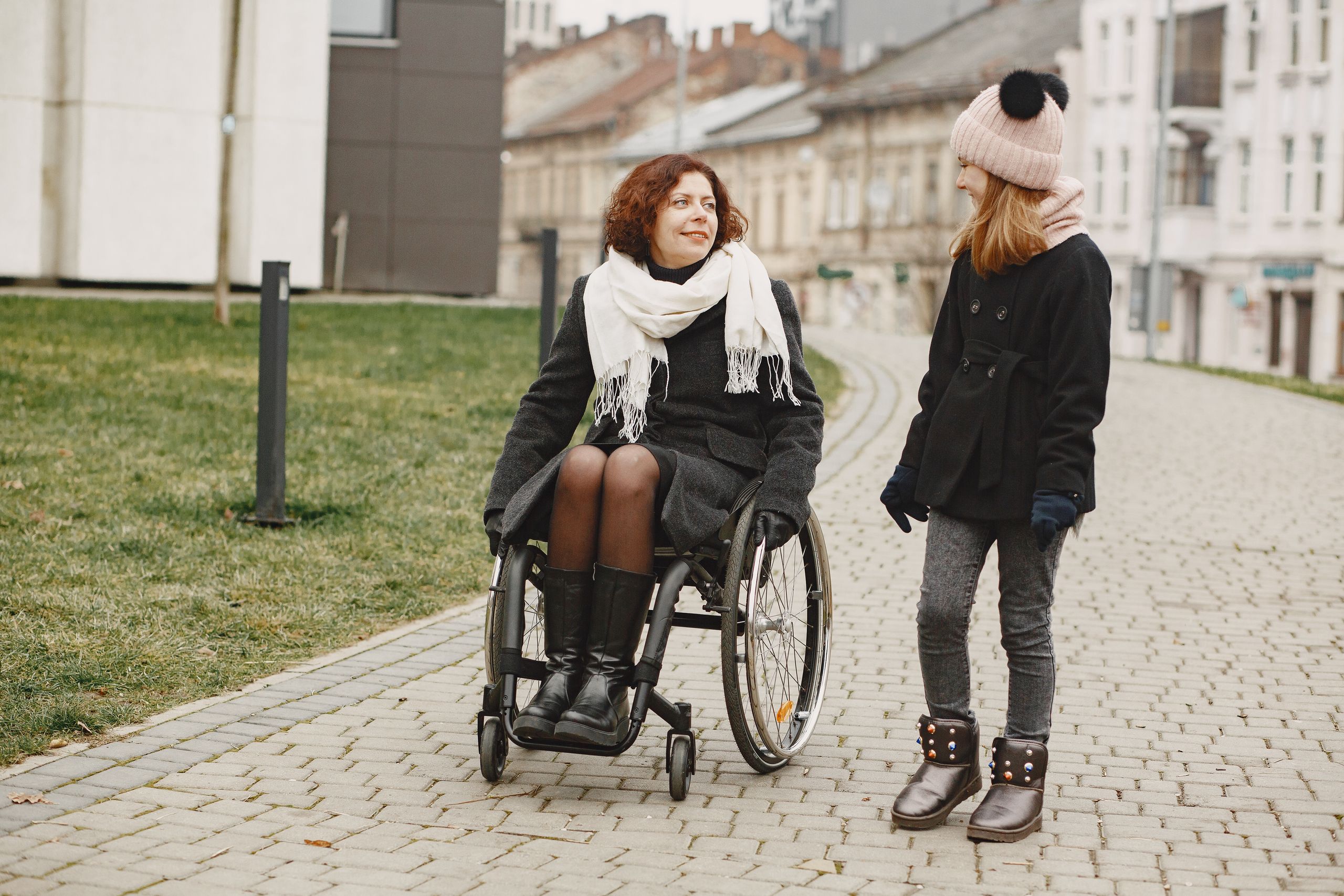 Woman in a wheelchair and a young girl walking on cobblestone path outdoors.